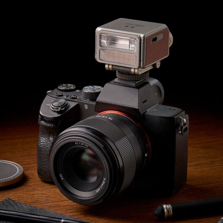 A black digital camera with a detachable lens and a rectangular external flash mounted on top, placed on a wooden surface with a lens cap and a pen nearby.