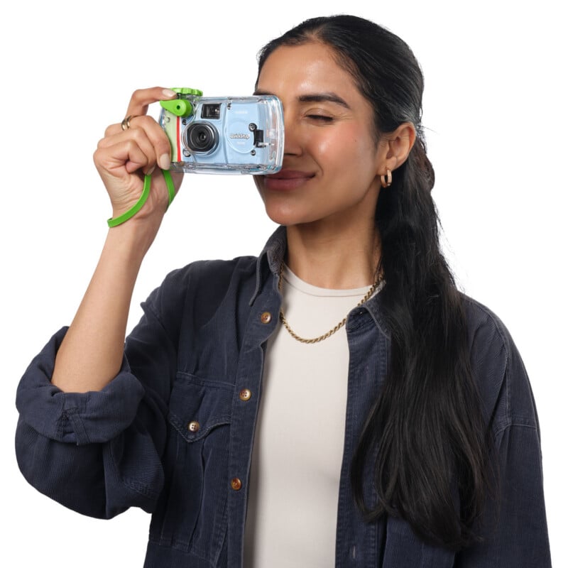 A woman with long dark hair, wearing a navy shirt and gold jewelry, smiles while holding a blue camera up to her eye as if taking a photo. She stands against a white background.