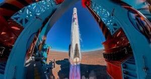 A rocket launches vertically from a launch pad, emitting bright flames and smoke, with metal support structures framing the view and a vast desert landscape in the background under a clear blue sky.