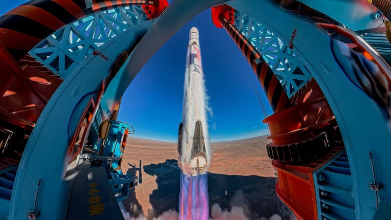 Un cohete se lanza verticalmente desde una plataforma de lanzamiento en el desierto, rodeado de estructuras de soporte azules y naranjas, con llamas brillantes y humo atravesando el cielo azul claro.