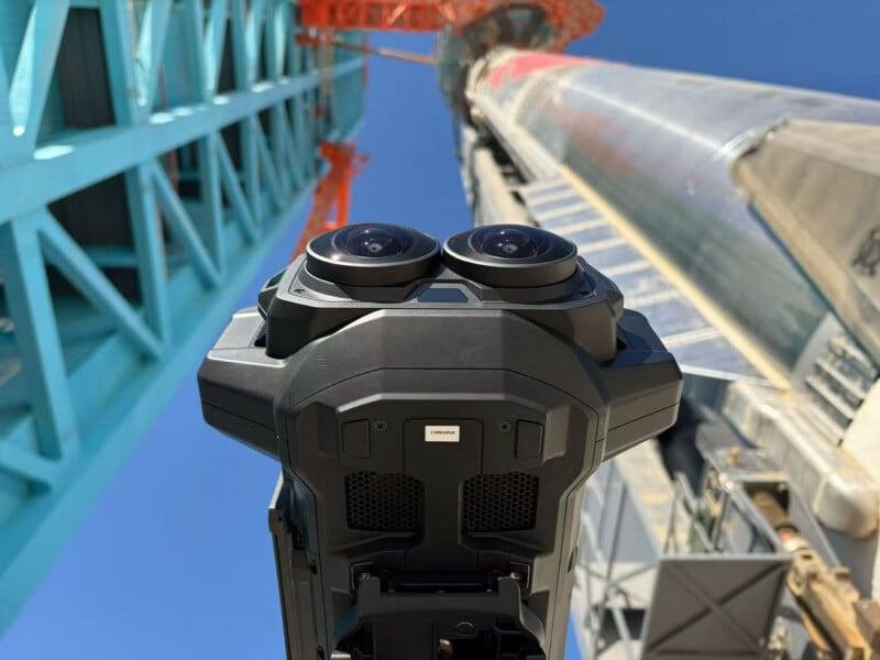 Primer plano de un dron negro con cámara de doble lente desde abajo, con una torre de lanzamiento y un cohete elevándose hacia el cielo azul claro en el fondo.