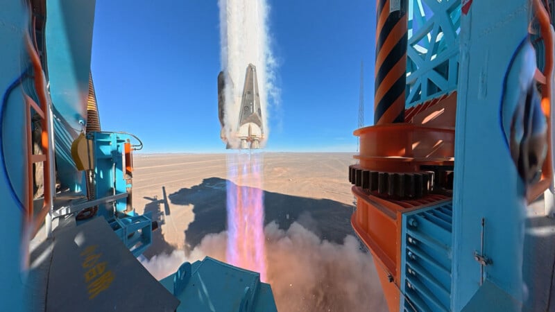 Una nave espacial se lanza verticalmente desde una plataforma en el desierto, emitiendo llamas brillantes y humo. El fondo es cielo azul y arena plana. El equipo estructural de lanzamiento se puede ver a ambos lados de la imagen.
