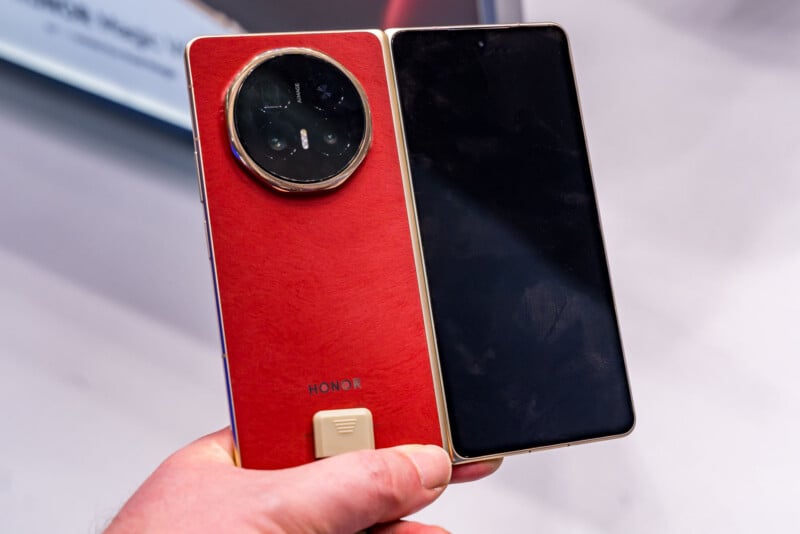 A hand holds an HONOR foldable smartphone in a half-open position, showing its large circular camera module and red back cover on the left, and its unfolded black screen on the right.