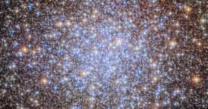 A dense cluster of bright blue, white, and orange stars against a dark background, resembling a sparkling, glowing patch in space. This image shows a globular star cluster with many stars close together.