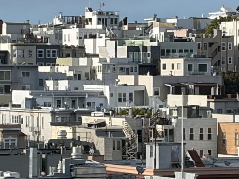 A dense cluster of multi-story apartment buildings and houses with flat roofs and balconies, closely packed together in an urban neighborhood, under a clear blue sky.