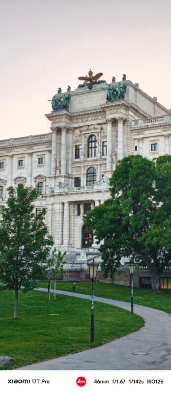 A grand historic building with ornate columns, statues, and an eagle sculpture on the roof stands behind a green lawn and trees, under a pale sky. Pathways and vintage street lamps are visible in the foreground.