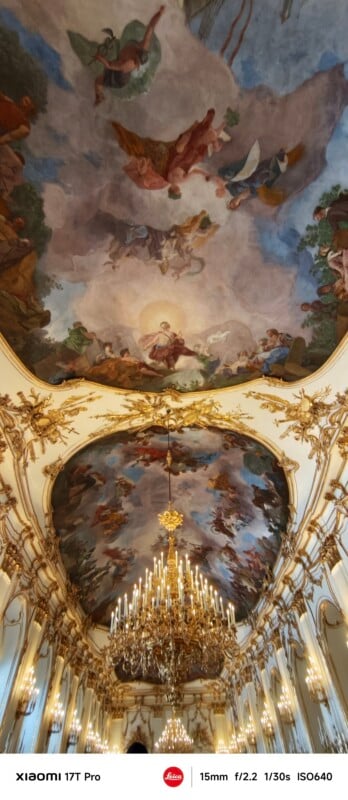 Ornate hall with golden chandeliers and elaborate Baroque ceiling frescoes depicting angels and figures in a heavenly scene, surrounded by decorative gold moldings and tall windows.