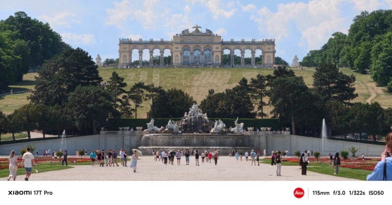 Visitors walk around a large fountain in a landscaped garden, with the Gloriette pavilion atop a hill in the background at Schönbrunn Palace, Vienna. Trees surround the area under a partly cloudy sky.
