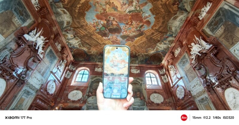 A hand holds a smartphone upward, displaying the ornate ceiling of a grand, baroque-style hall with intricate frescoes, columns, and large windows, aligning the phone’s view with the real ceiling above.