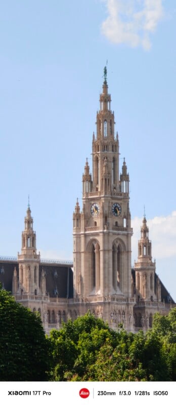 A grand neo-Gothic building with tall spires and a clock tower rises above green trees under a clear blue sky. Camera settings are visible at the bottom of the image.