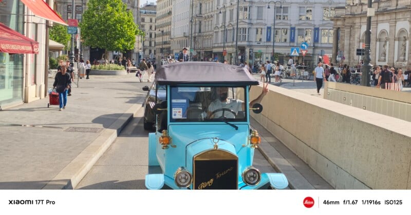 A vintage blue car drives down a city street lined with pedestrians, shops, and historic buildings on a sunny day. The driver is visible through the windshield. Camera settings are displayed at the bottom of the image.