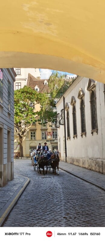 A horse-drawn carriage with passengers travels down a cobblestone street lined with historic buildings under a yellow archway on a sunny day.