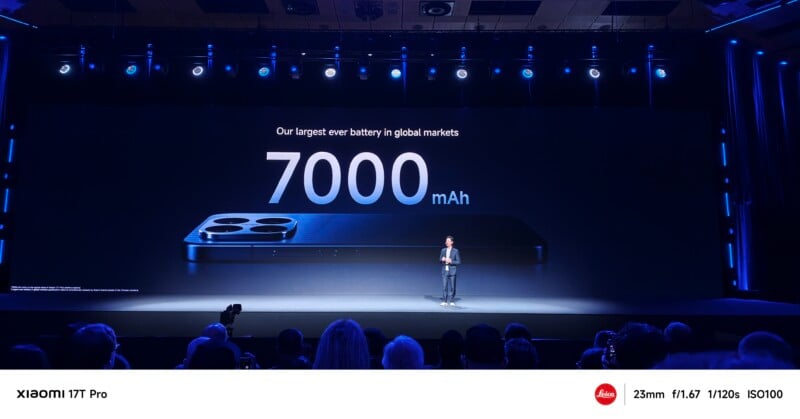 A presenter stands onstage in front of a large screen displaying “7000 mAh” and an image of a smartphone, announcing the battery capacity to an audience at a technology event.