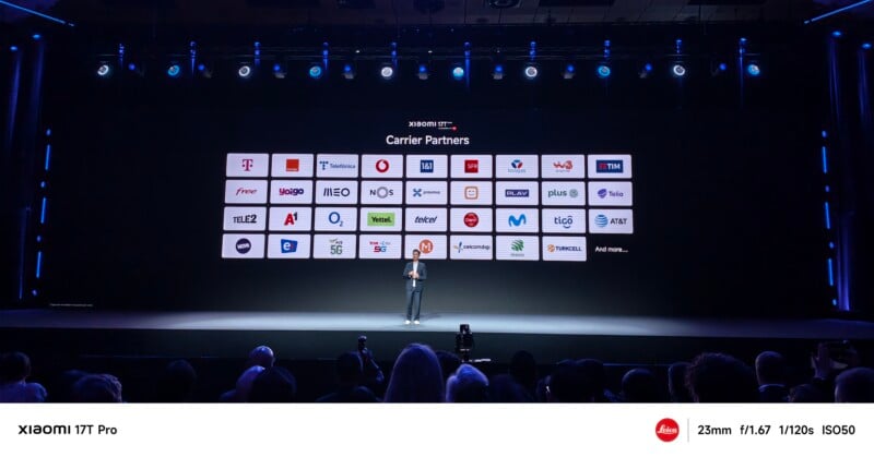 A presenter stands on stage in front of a large screen displaying the logos of various carrier partners at a technology event. The audience watches from the foreground.