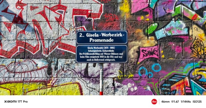 A blue street sign on a graffiti-covered wall reads "2. Gisela-Werbezirk-Promenade," honoring an actress and mentioning her Hollywood success. The colorful graffiti includes various abstract shapes and tags.