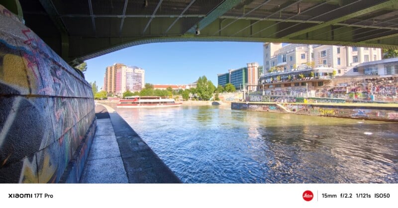 View from under a bridge showing a river with a sightseeing boat, buildings, and graffiti-covered walls on a sunny day. Camera specs are shown at the bottom of the image.