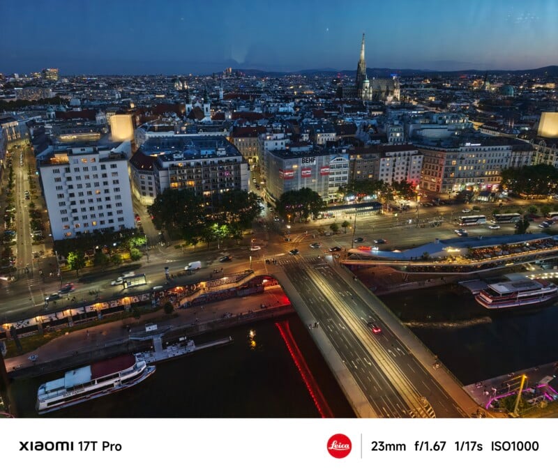 Aerial view of a cityscape at dusk, featuring a river crossed by a busy bridge, illuminated buildings, bustling streets with cars, and distant city lights under a darkening blue sky. Camera info overlay at the bottom.