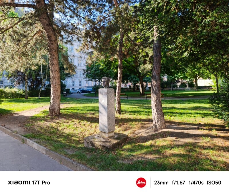A small stone pedestal with a bust stands among trees and grass in a city park on a sunny day. A path runs nearby, and a white building is visible in the background.