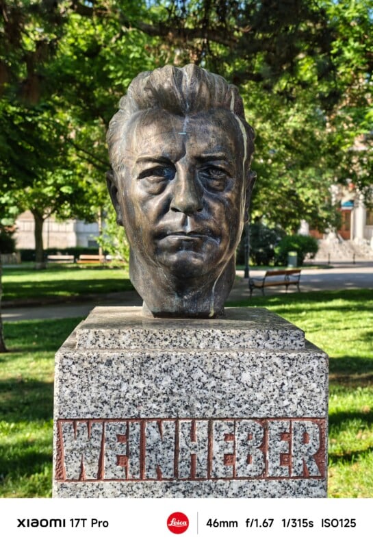 Bronze bust of a man on a granite pedestal inscribed with "WEINHEBER," set in a sunlit park with green trees and a bench in the background.