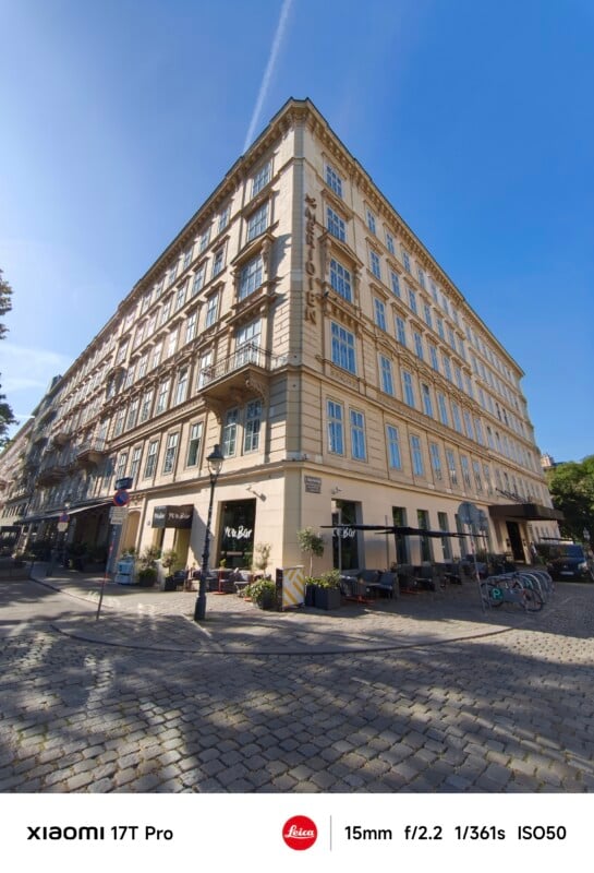 A wide-angle view of a corner building with cream-colored walls and many windows, located on a cobblestone street under a clear blue sky. Bicycles and a few sidewalk cafes are visible at street level.