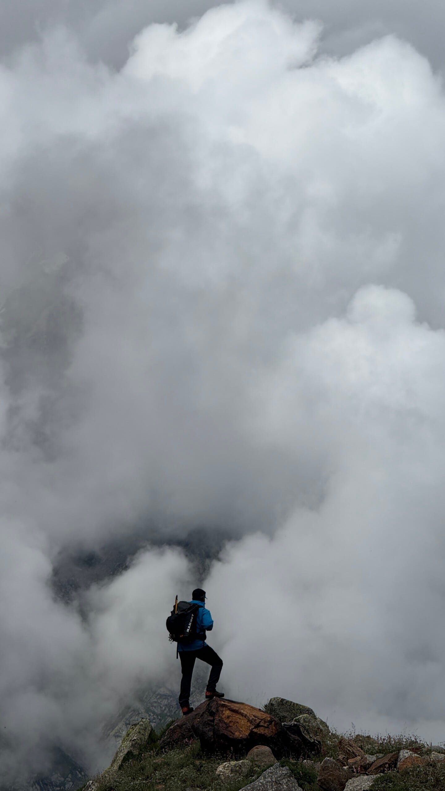 A person wearing a backpack stands on a rocky ledge, gazing into thick, billowing clouds that obscure the landscape ahead, creating a dramatic and adventurous atmosphere.
