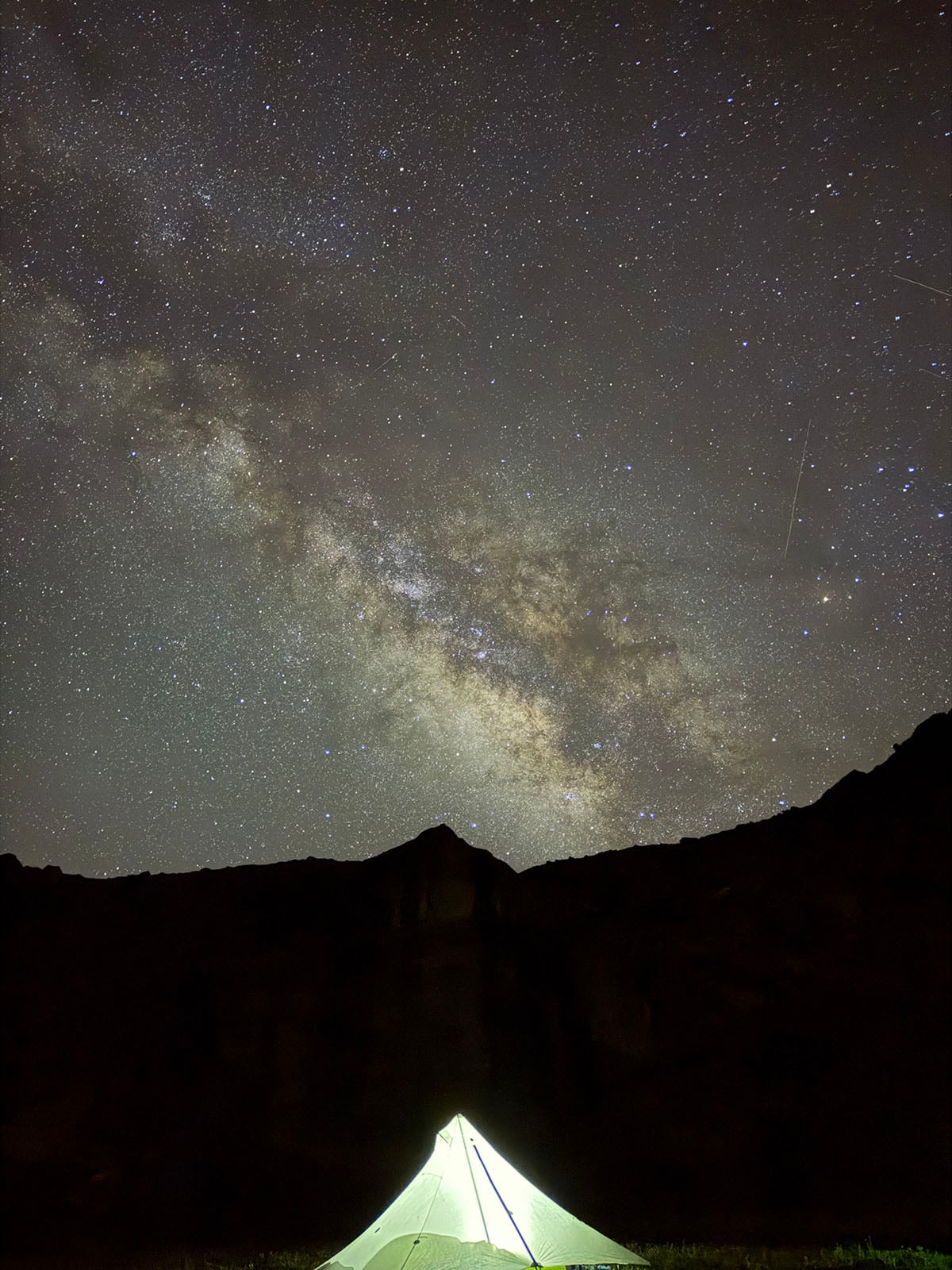 A glowing tent sits on the ground beneath a dark, star-filled sky with the Milky Way galaxy prominently visible above silhouetted mountains.