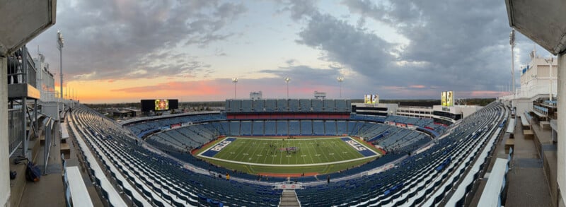 A wide panoramic view of an empty football stadium at sunset, with blue seats, a green field marked "BILLS," and a colorful sky with clouds in the background.