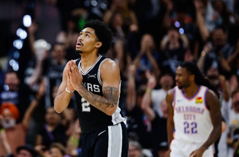 A basketball player in a black Spurs jersey stands with hands clasped and looks upward during a game, while a player in a white Oklahoma City jersey walks in the background and the crowd watches.