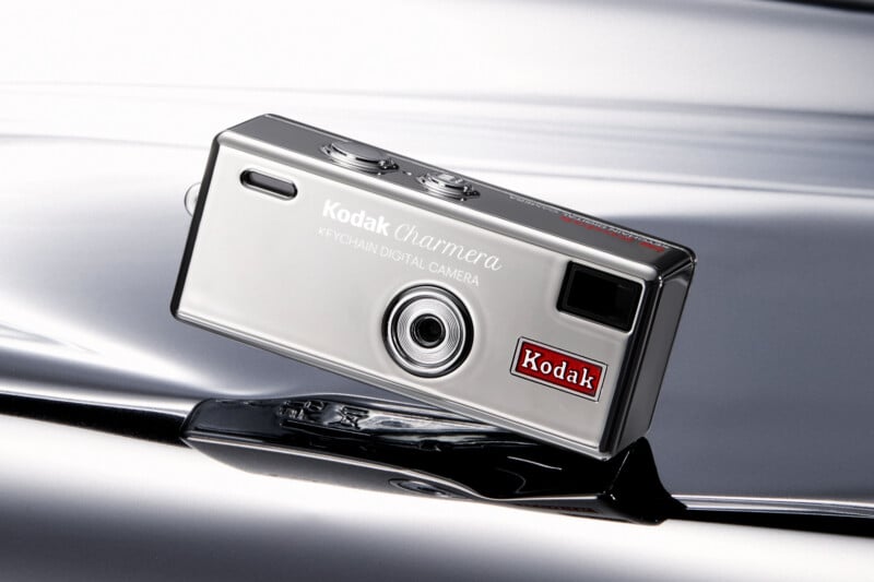 A small, rectangular silver Kodak keychain digital camera is displayed against a reflective metallic surface, with the Kodak logo visible on the front.