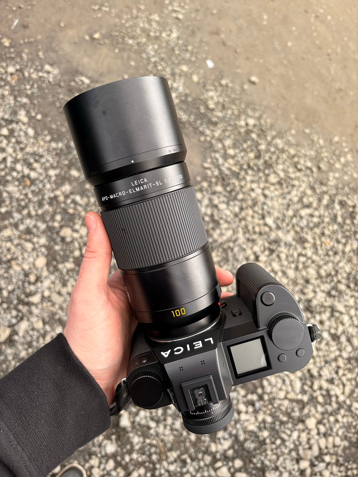 A hand holds a Leica camera with a large Leica APO-Macro-Elmarit-SL 100mm lens, positioned over a ground covered in small rocks and gravel.