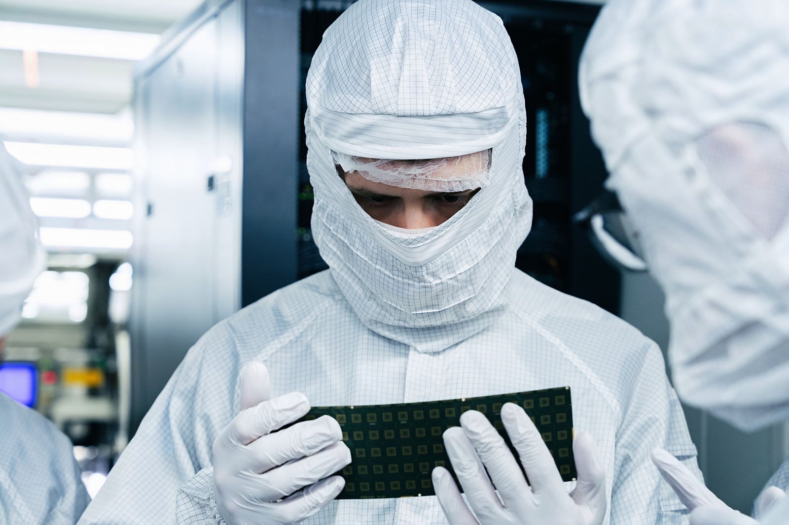 A person in a cleanroom suit, gloves, and face mask examines a green rectangular electronic component, with others in similar attire nearby, inside a high-tech laboratory or manufacturing facility.