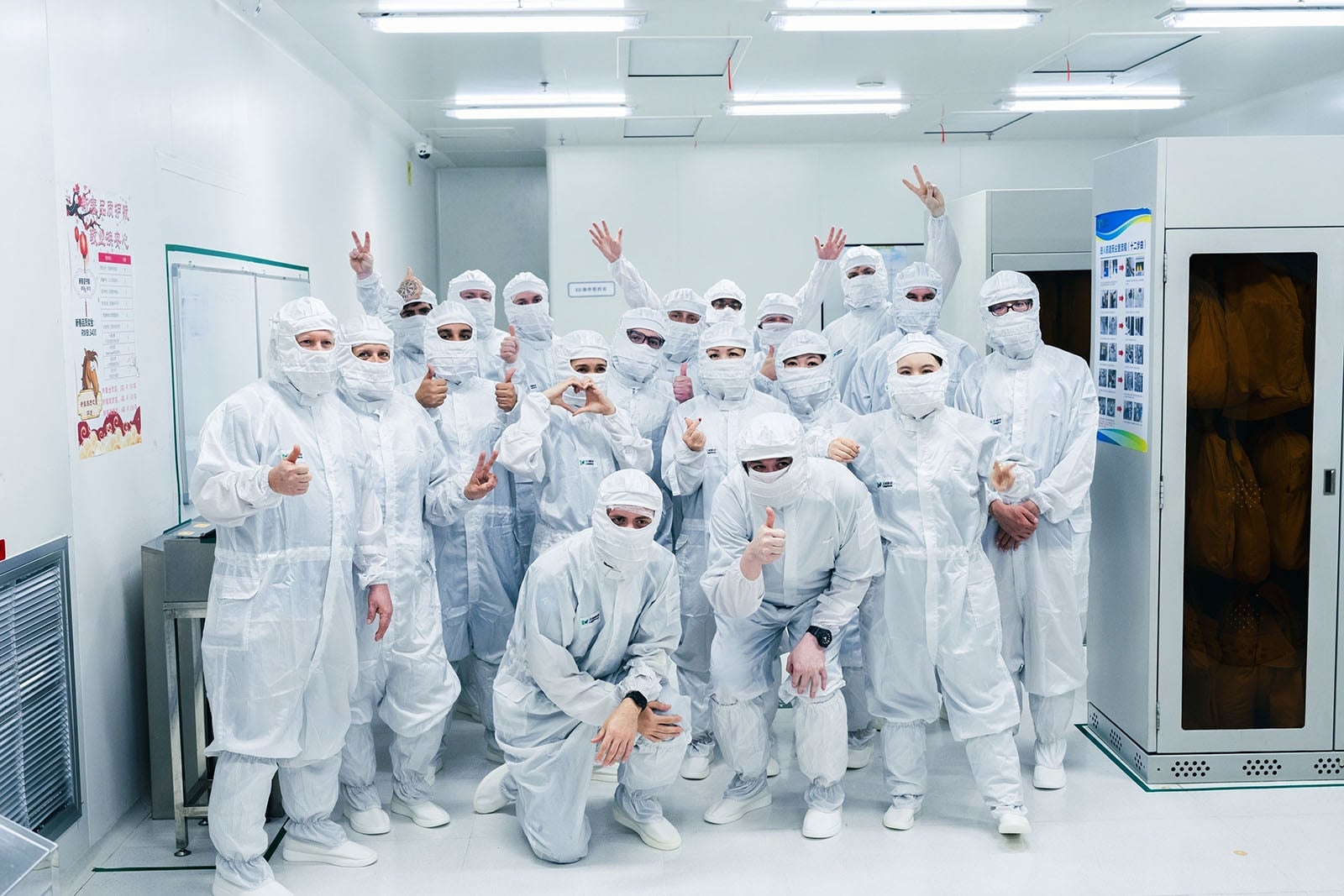 A group of people wearing full-body white protective suits, masks, and hair covers pose together in a clean, brightly lit room, making peace signs and thumbs-up gestures.