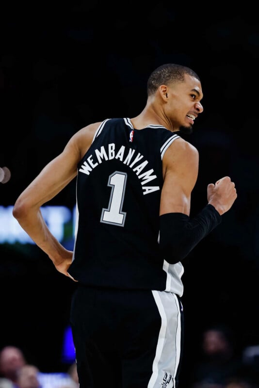 A basketball player in a black jersey with "Wembanyama" and the number 1 on the back raises his arm and looks over his shoulder during a game.
