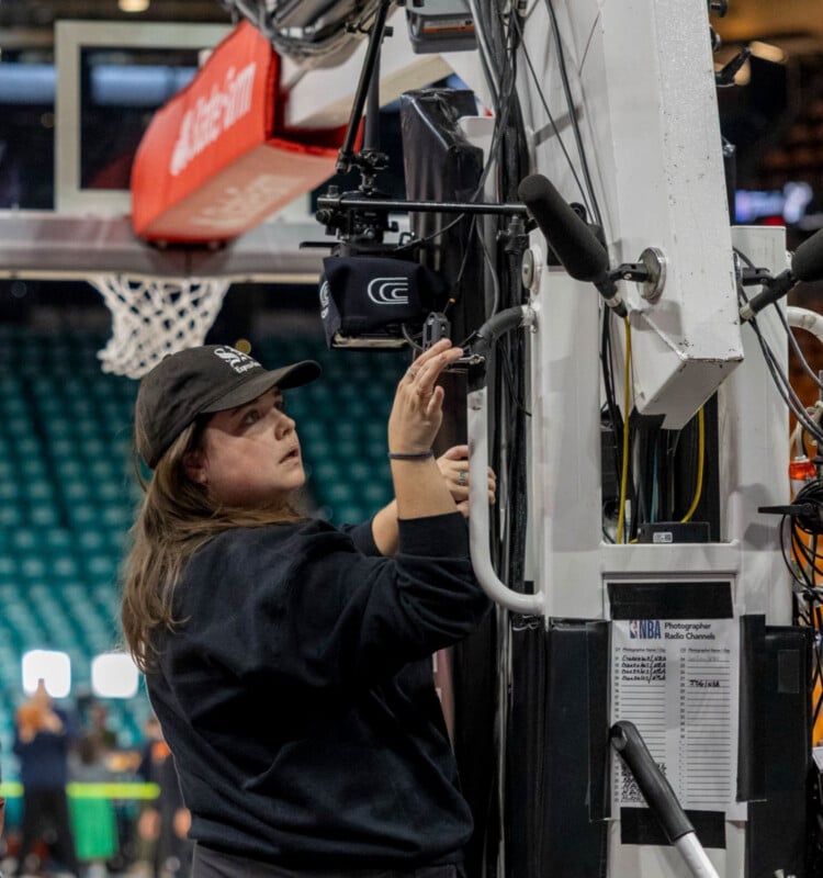 A woman in a black sweatshirt and cap adjusts camera equipment attached to a basketball hoop support on a court, with the net and an audience blurred in the background.