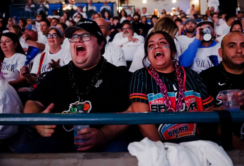 Two excited fans wearing San Antonio Spurs gear cheer loudly in a crowded stadium, surrounded by other enthusiastic spectators. The lively atmosphere is filled with people wearing team colors and accessories.