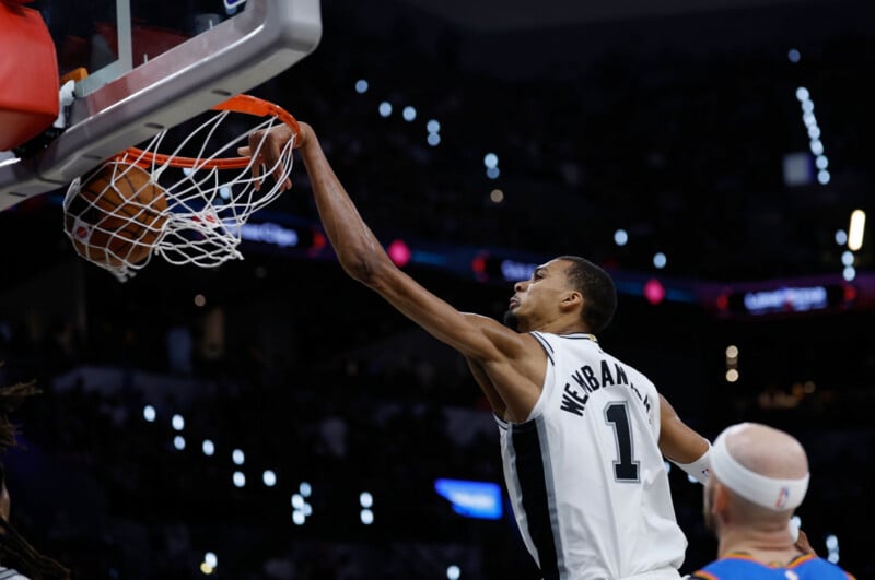 A basketball player in a Spurs jersey, number 1, stretches to dunk the ball into the hoop during a game, with an opposing player in a white headband watching nearby.