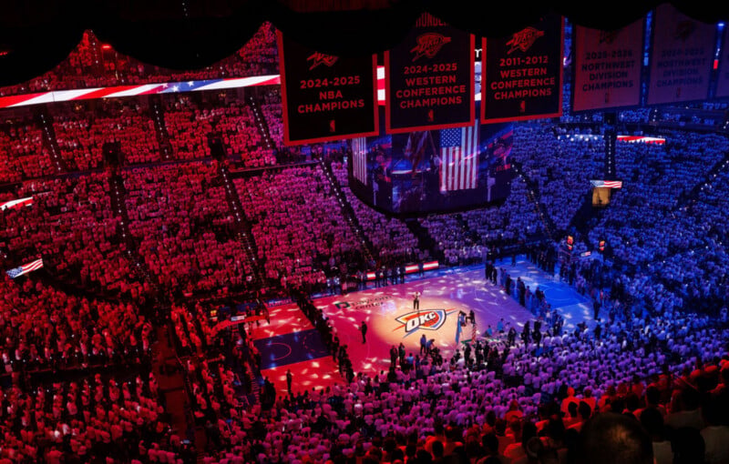 A packed arena with fans in red and blue lighting surrounds a basketball court with the Oklahoma City Thunder logo; championship banners hang above the court.