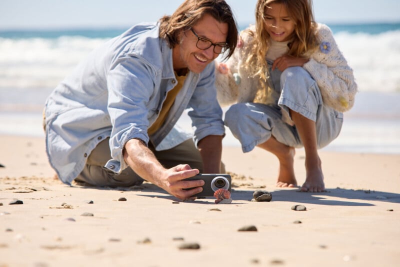 A man and a young girl crouch on a sandy beach, smiling as the man takes a photo of seashells and rocks with a camera. Waves and blue sky are visible in the background.