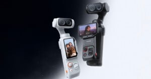 Two handheld 3D cameras, one white and one black, each with a touchscreen displaying selfies, stand side by side against a gradient black-to-white background. Both have dual lenses and control buttons.