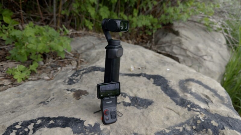 A handheld gimbal camera with a small screen and control buttons is placed upright on a large rock outdoors, surrounded by green plants and foliage.