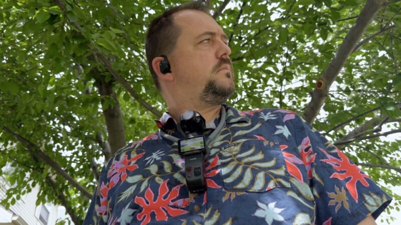A man wearing a colorful, floral-patterned shirt stands outdoors under leafy green trees, looking to his left. He has a beard, wears a wireless earpiece, and has devices attached to a strap on his chest.