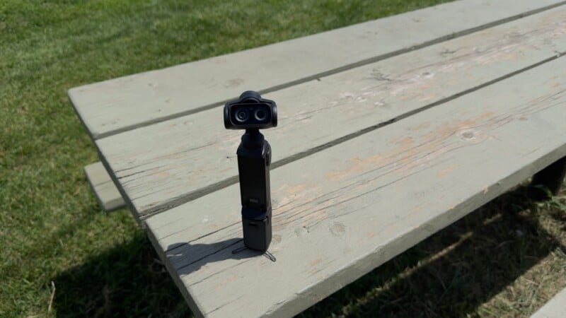 A small handheld camera with a gimbal stands upright on a weathered wooden picnic table, with green grass visible in the background.