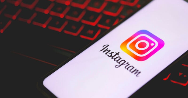 A smartphone displaying the Instagram logo and name rests on a surface, with a red-lit keyboard blurred in the background.
