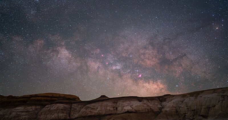A star-filled night sky with the Milky Way galaxy glowing above rugged, rocky cliffs in the foreground. The scene highlights bright stars, cosmic dust, and soft, colorful nebulae stretching across the horizon.