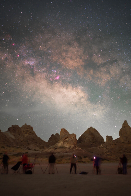 People with tripods and cameras stand in a desert landscape at night, photographing a brightly lit, star-filled sky and the Milky Way above jagged rocky formations.