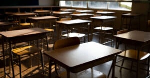 Rows of empty wooden desks and chairs in a classroom, lit by sunlight streaming through large windows. The room is quiet and unoccupied, with shadows cast across the floor and desks.