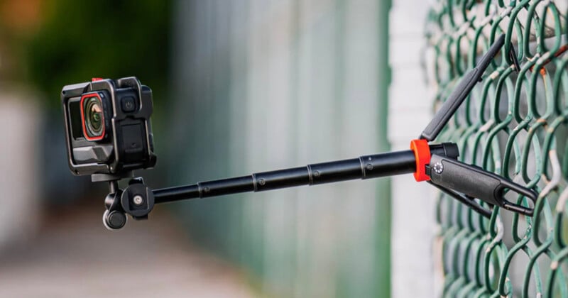 A black action camera mounted on a telescopic pole is clamped to a green chain-link fence, with the camera pointing outward, ready to capture footage.