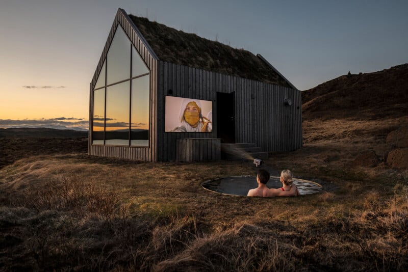 A couple relaxes in a hot tub outside a modern cabin with large glass windows and a grass roof, watching a movie projected on the cabin’s wall at sunset in a rugged, open landscape.