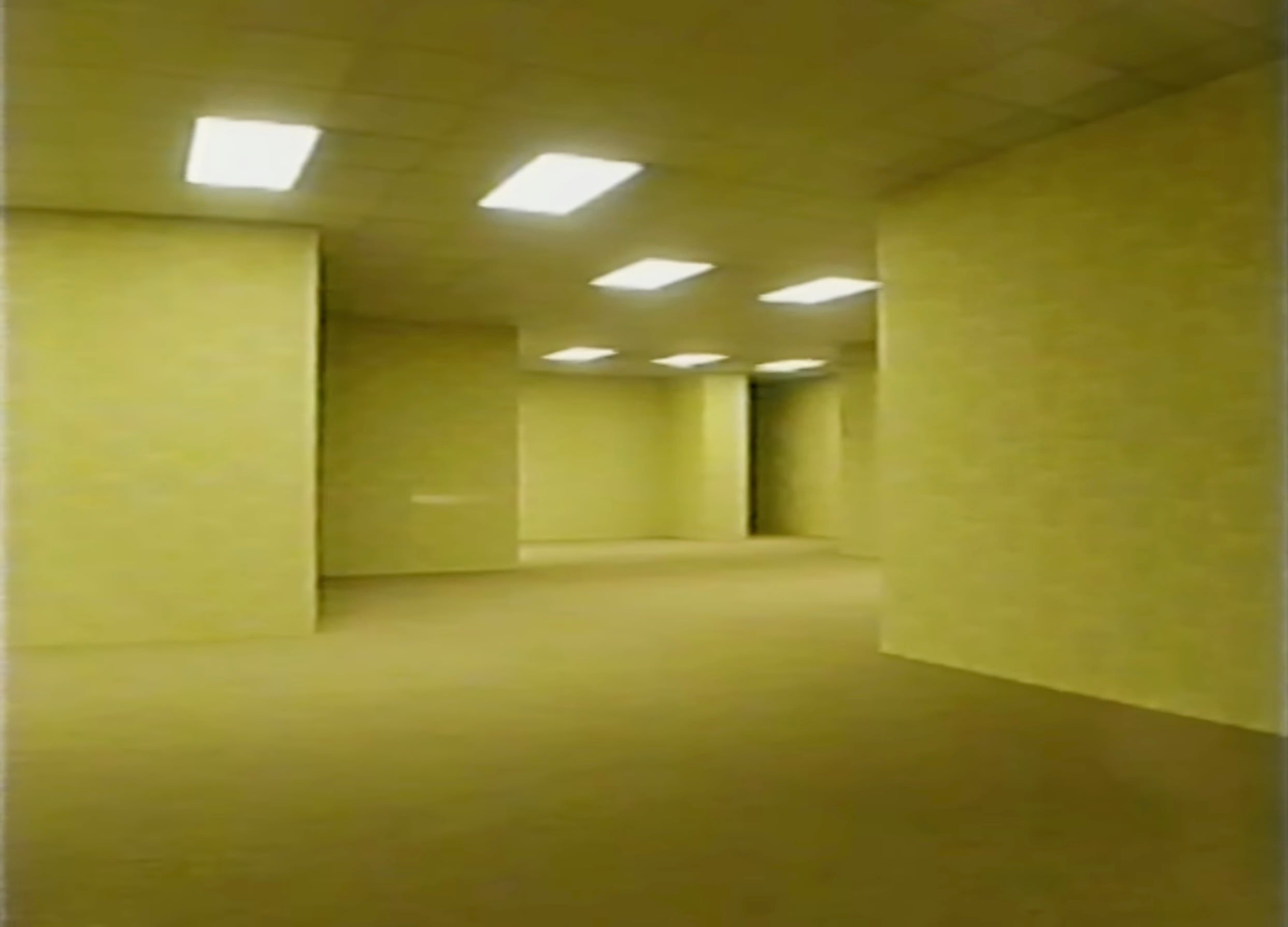 A dimly lit, empty room with yellowish walls, a patterned carpet, and fluorescent ceiling lights, creating a maze-like, eerie atmosphere. There are no windows or furniture visible.