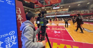 A cameraman films a basketball game from the sidelines inside an indoor arena, where players are warming up on the court and spectators are seated in the stands.